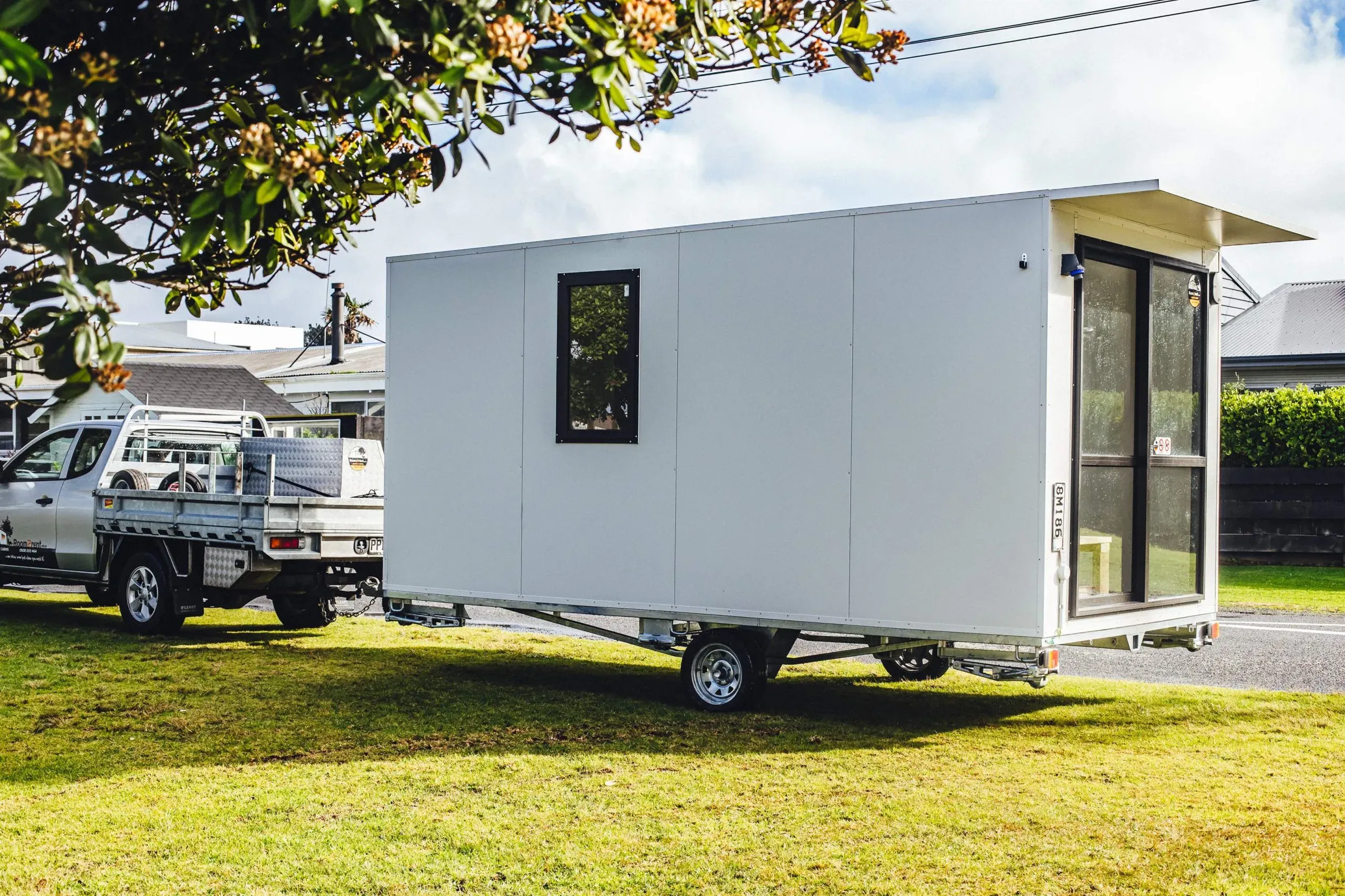 A tiny white mobile home is parked on the grass, blending into the natural landscape around it