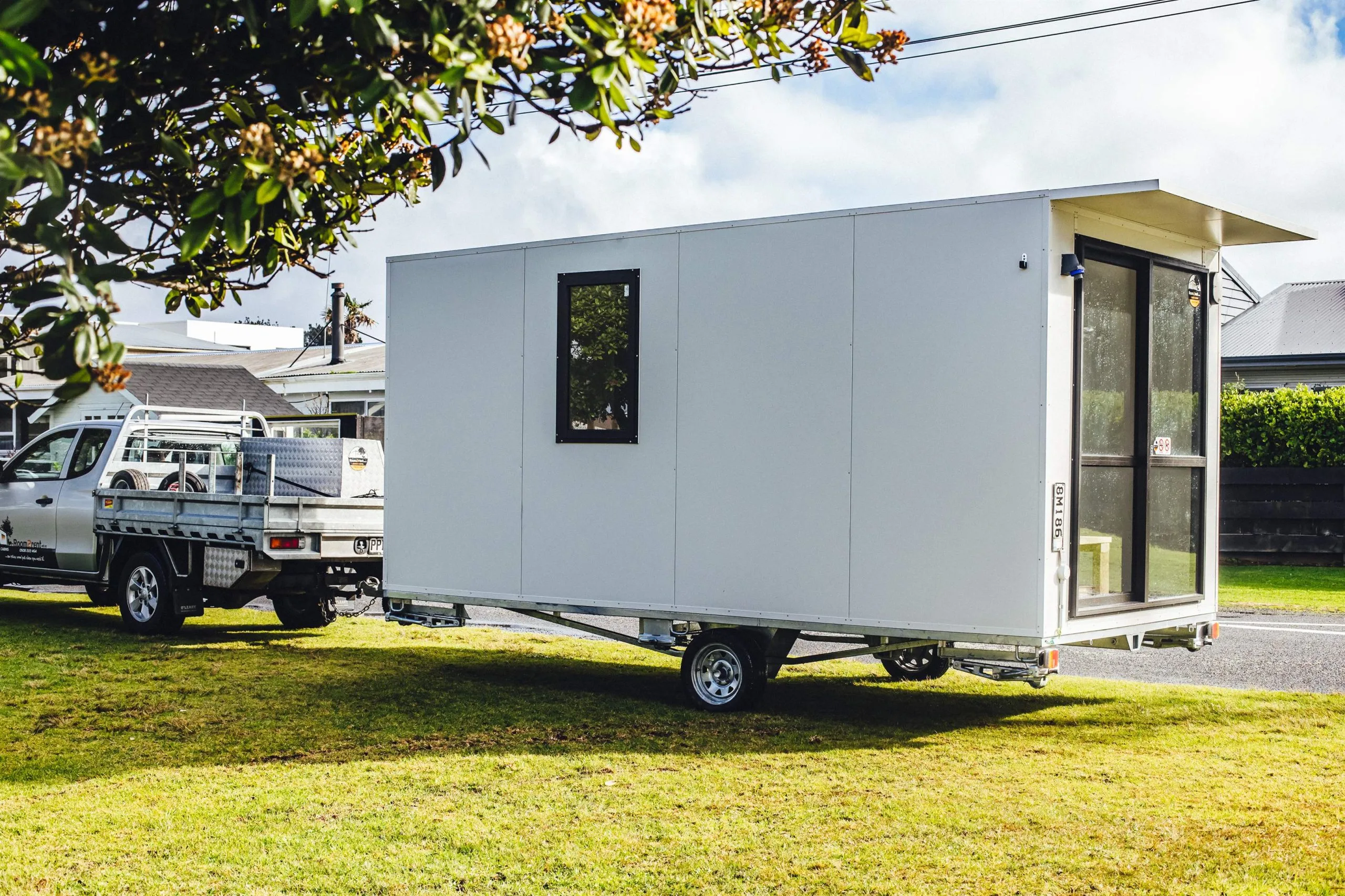 A tiny white mobile home is parked on the grass, blending into the natural landscape around it