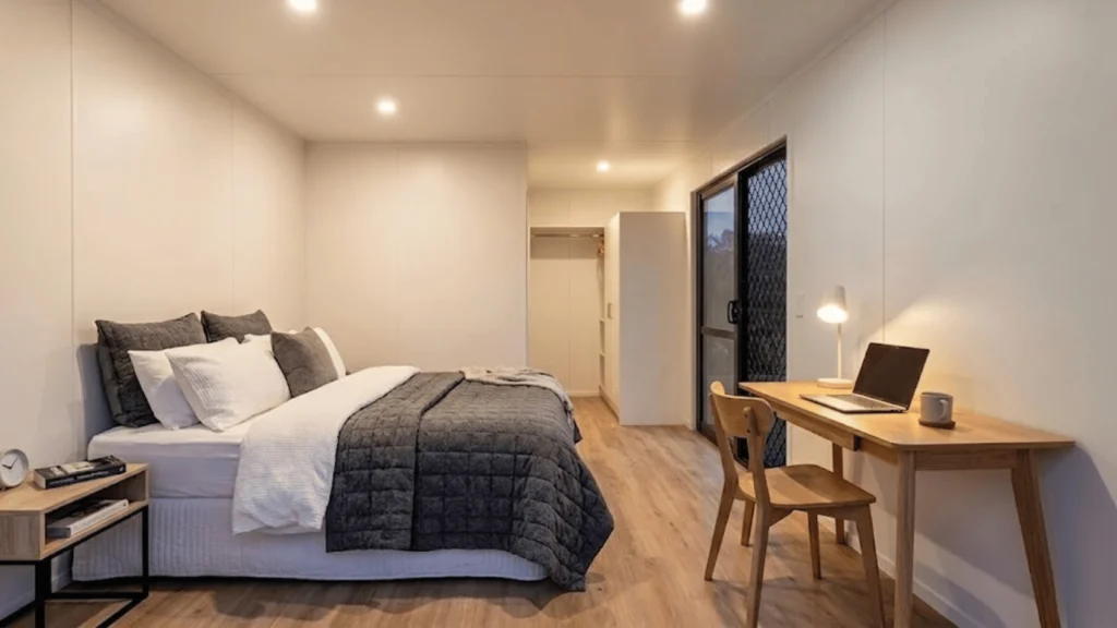 On the left, a queen-sized bed with thick white and dark grey winter bedding. On the right, a neat timber desk with a laptop and a warm table lamp. The walls are a smooth, clean white. cabins to rent rotorua