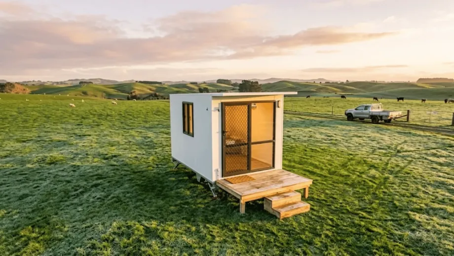 The portable cabins waikato has a small wooden deck and a glowing warm light shining through the window. Frost is lightly visible on the grass.